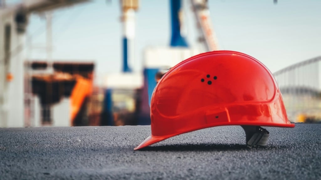 A red hard hat lying on the ground at a construction site, symbolizing workplace safety and emergency management preparedness. The blurred background of industrial equipment highlights the importance of raising the standard in occupational safety in Hawaii.